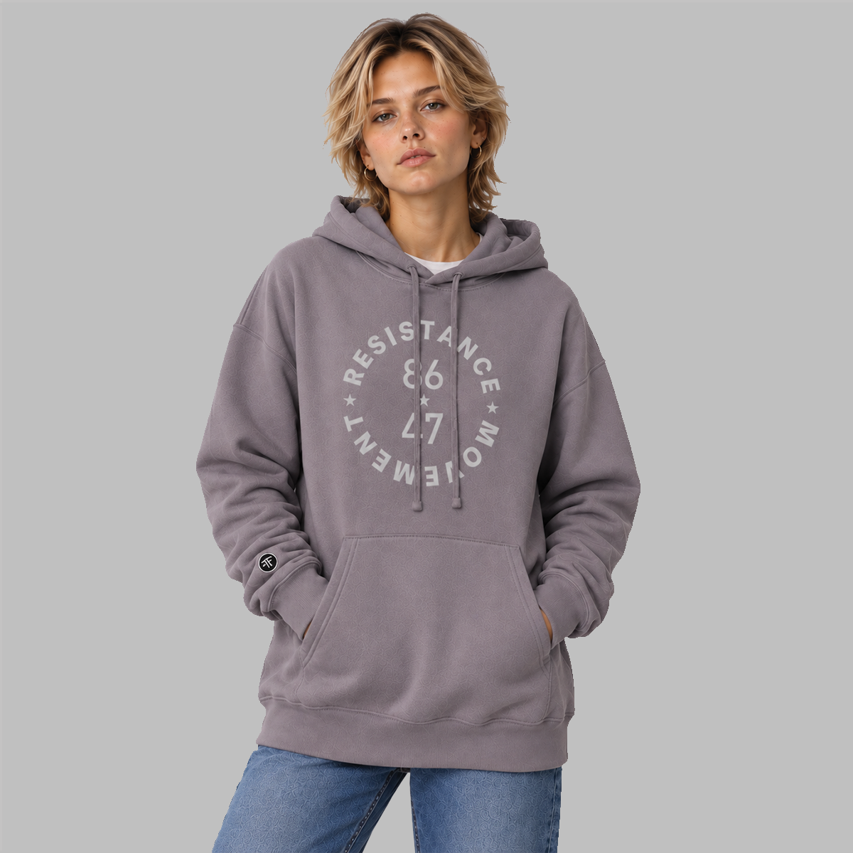 A woman wearing a slate purple pullover hoodie with a light grey circular graphic on the chest that reads 'RESISTANCE MOVEMENT 86 47' and blue jeans, standing against a solid gray studio background.