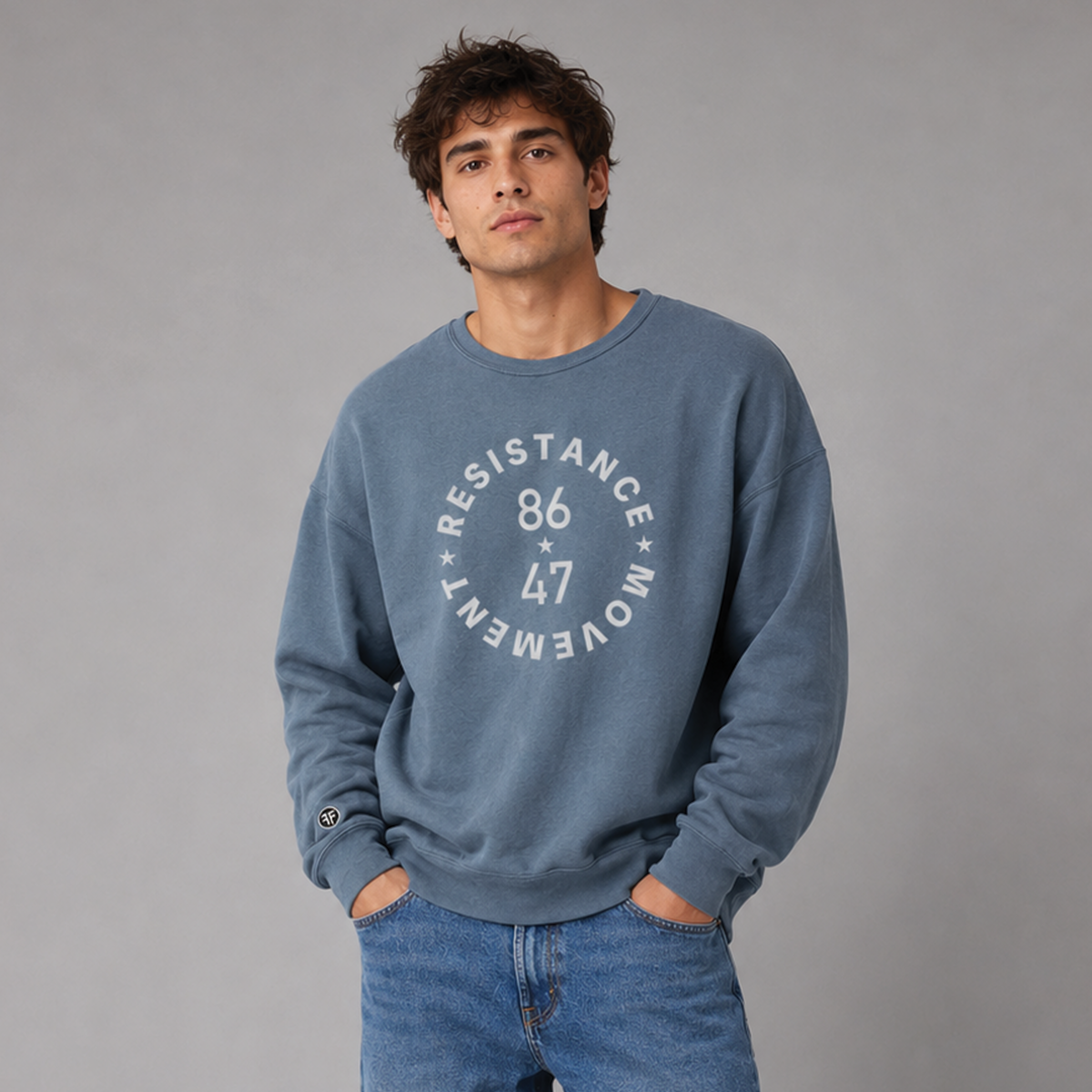 A man wearing a dusty blue crewneck sweatshirt with a light grey circular graphic that reads 'RESISTANCE MOVEMENT 86 47' on the chest, paired with blue jeans, standing against a solid gray studio background.