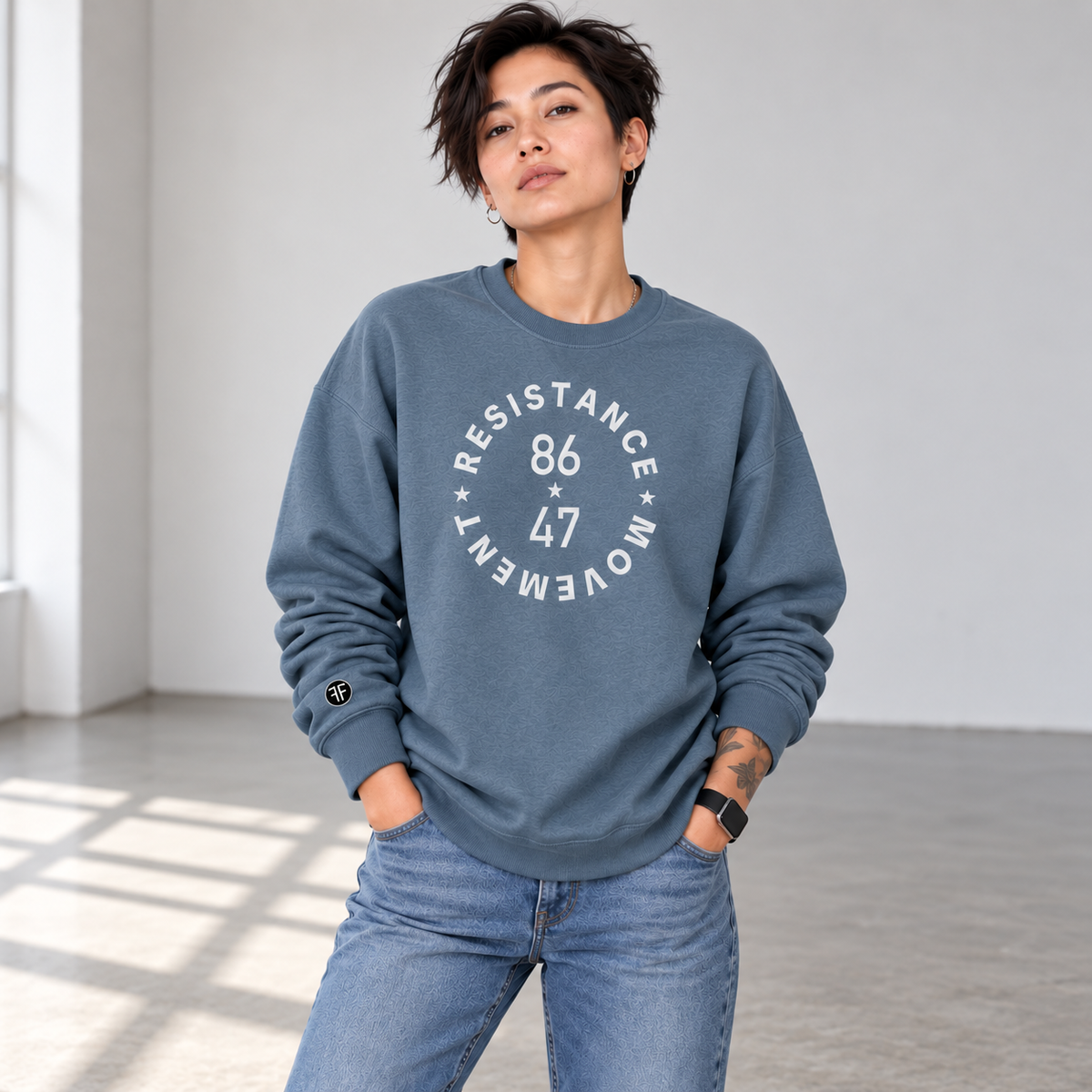 A female model wearing a slate blue crewneck sweatshirt with a light grey circular graphic that reads 'RESISTANCE MOVEMENT 86 47' on the front, paired with blue jeans in a bright, modern studio setting.