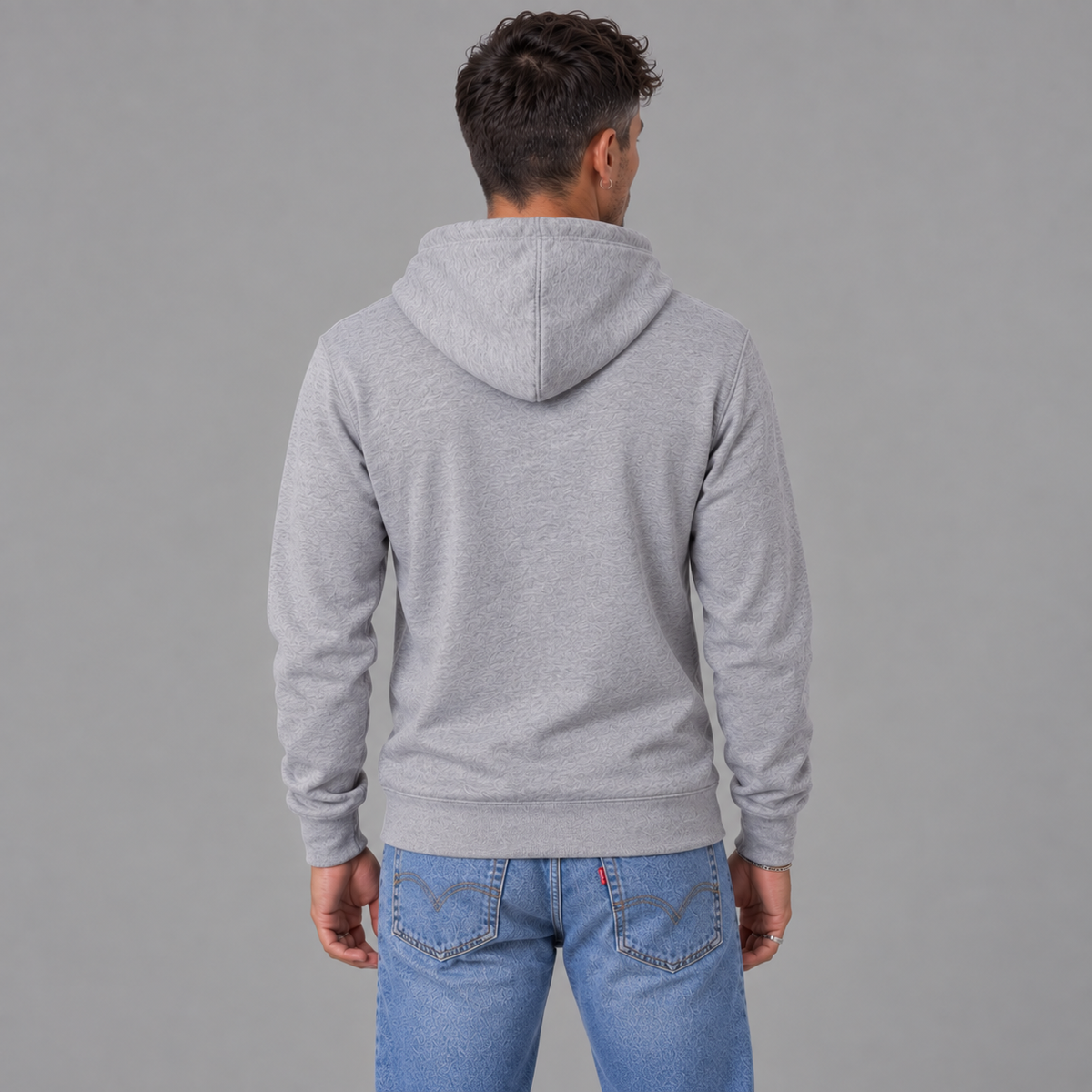 A man viewed from behind wearing a light gray heather pullover hoodie and blue jeans, standing against a solid gray studio background to show there are graphics on the back.