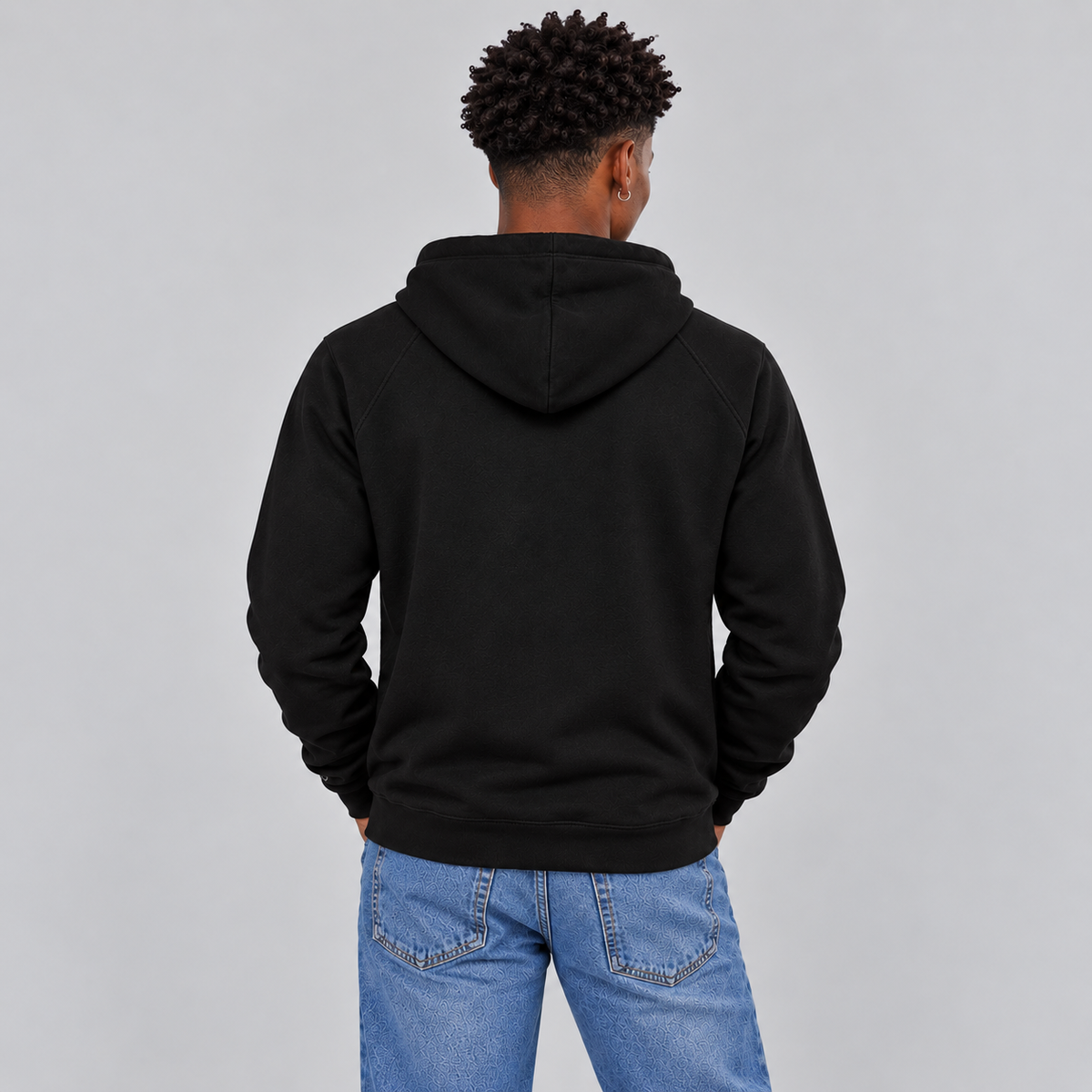 A man viewed from behind wearing a black pullover hoodie and blue jeans, standing against a solid light gray studio background.