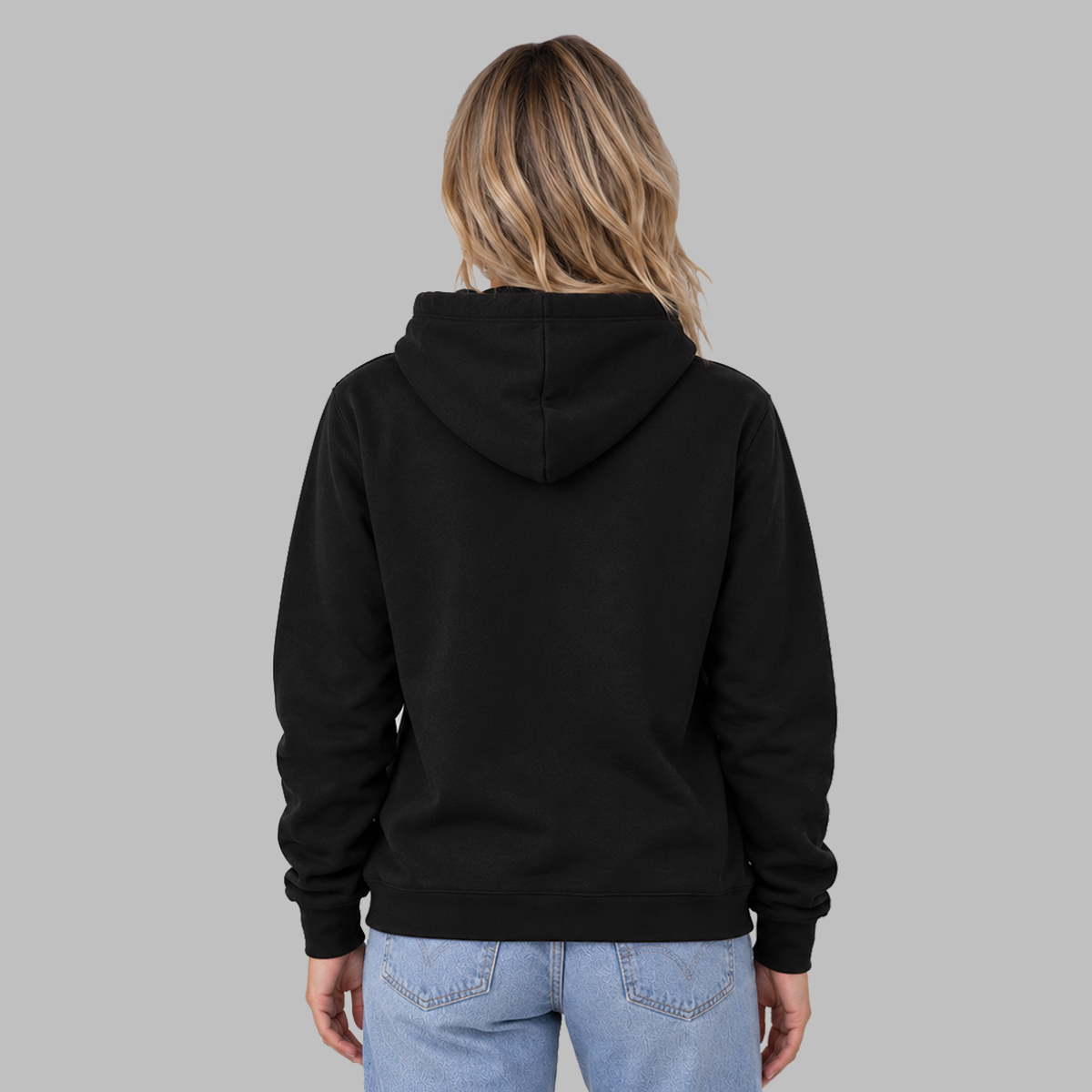 A woman viewed from the back wearing a plain black pullover hoodie and light blue jeans, standing against a solid light gray studio background.