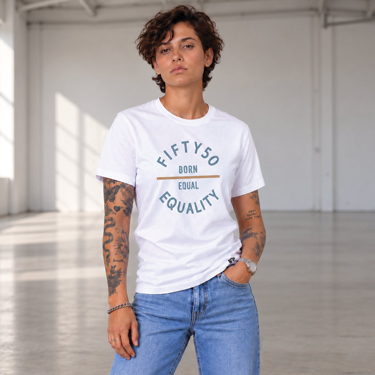 A woman with short dark hair and tattoos stands in a bright industrial loft wearing a white graphic T-shirt and light blue jeans. The shirt has a circular slate blue and soft orange graphic that reads 'FIFTY50 BORN EQUAL EQUALITY'.