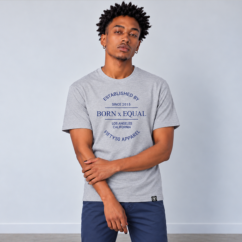 A man wearing a heather gray graphic t-shirt with navy blue text that reads 'BORN X EQUAL' and 'ESTABLISHED BY FIFTY50 APPAREL', paired with navy blue chinos, standing against a light gray wall.