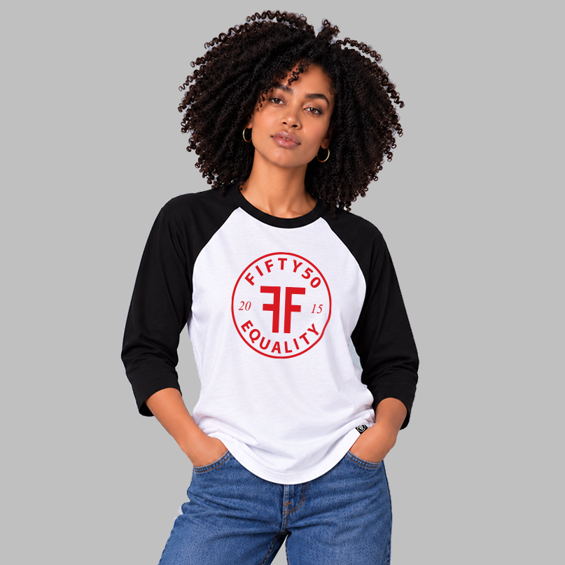 A woman with curly black hair wearing a white and black raglan baseball t-shirt with a red circular 'FIFTY50 EQUALITY' graphic on the chest, paired with blue jeans, against a solid gray background.