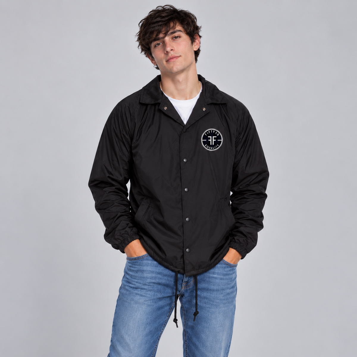 A man wearing a black lightweight coach's jacket with a circular black patch left chest that has two FFs in the middle, surrounded by 'Fifty50 Apparel' and 'Equality'. The jacket is paired with a white T-shirt and light blue jeans.