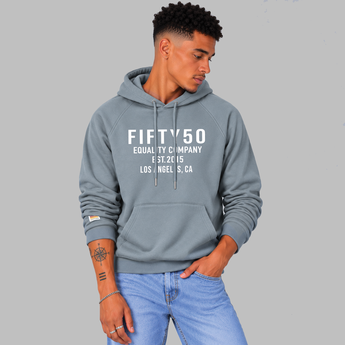 A man wearing a slate blue pullover hoodie with white text reading 'FIFTY50 EQUALITY COMPANY EST 2015 LOS ANGELES, CA' on the chest, paired with light blue jeans, against a solid gray studio background.