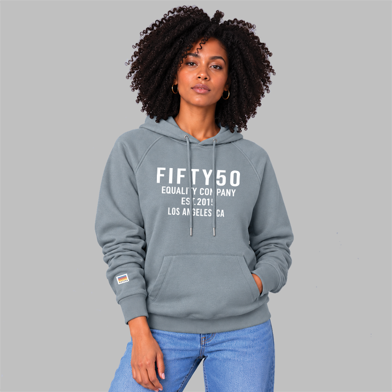 A woman wearing a slate blue pullover hoodie with white text reading 'FIFTY50 EQUALITY COMPANY EST 2015 LOS ANGELES, CA' on the chest, paired with light blue jeans, against a solid gray studio background.