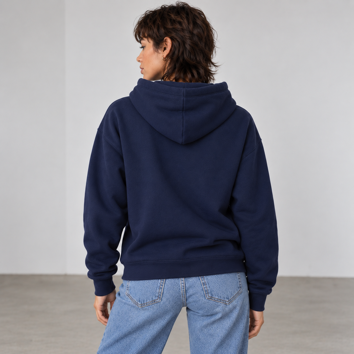 A woman shown from the back wearing a navy blue pullover hoodie showing the fit and that there is no logos on the back in a minimalist studio setting with a light gray wall and concrete floor.