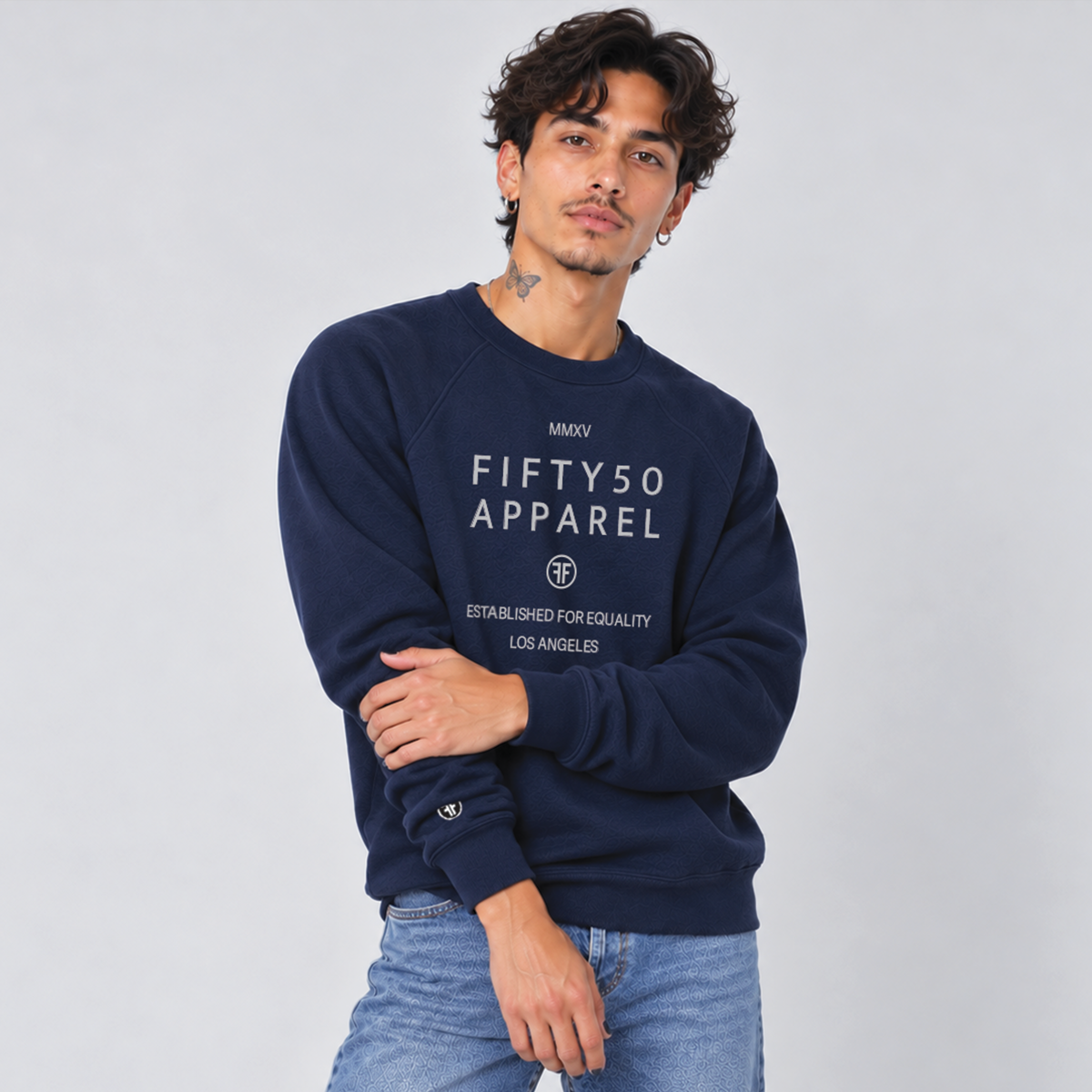 A man wearing a navy blue crewneck sweatshirt with 'FIFTY50 APPAREL ESTABLISHED FOR EQUALITY LOS ANGELES' printed in light grey on the front, paired with light-wash blue jeans, against a light gray studio background.