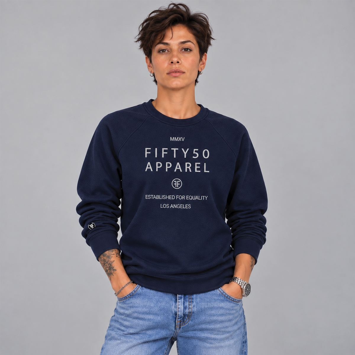 A woman with short hair wearing a navy blue crewneck sweatshirt with a light grey printed text reading 'FIFTY50 APPAREL ESTABLISHED FOR EQUALITY LOS ANGELES' paired with light wash blue jeans, against a solid gray background.