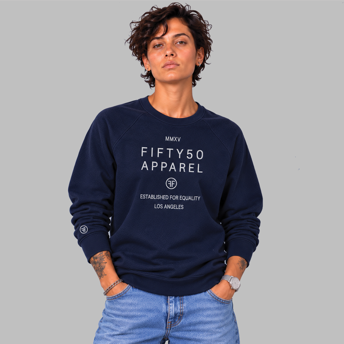 A woman wearing a navy blue crewneck sweatshirt with 'FIFTY50 APPAREL ESTABLISHED FOR EQUALITY LOS ANGELES' printed in light grey on the front, paired with light-wash blue jeans, against a light gray studio background.