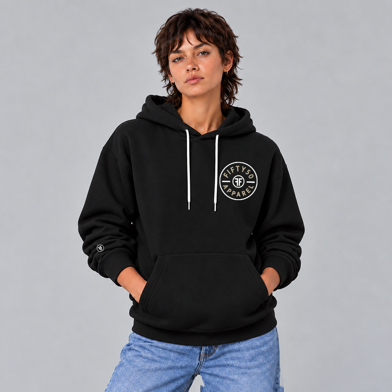 A woman wearing a black pullover hoodie with a circular white chest graphic that reads 'FIFTY50 APPAREL' in tan with two Fs back to back in white in the center. There is a small black and white Fifty50 logo on the sleeve.