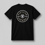 A black short-sleeve t-shirt with a circular white chest graphic that reads 'FIFTY50 APPAREL' with two white FFs back to back in the middle, paired with blue jeans, set against a solid gray background.