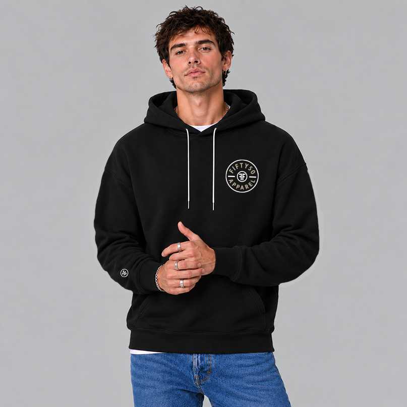 A man wearing a black pullover hoodie with a circular white chest graphic that reads 'FIFTY50 APPAREL' in tan with two Fs back to back in white in the center. There is a small black and white Fifty50 logo on the sleeve.