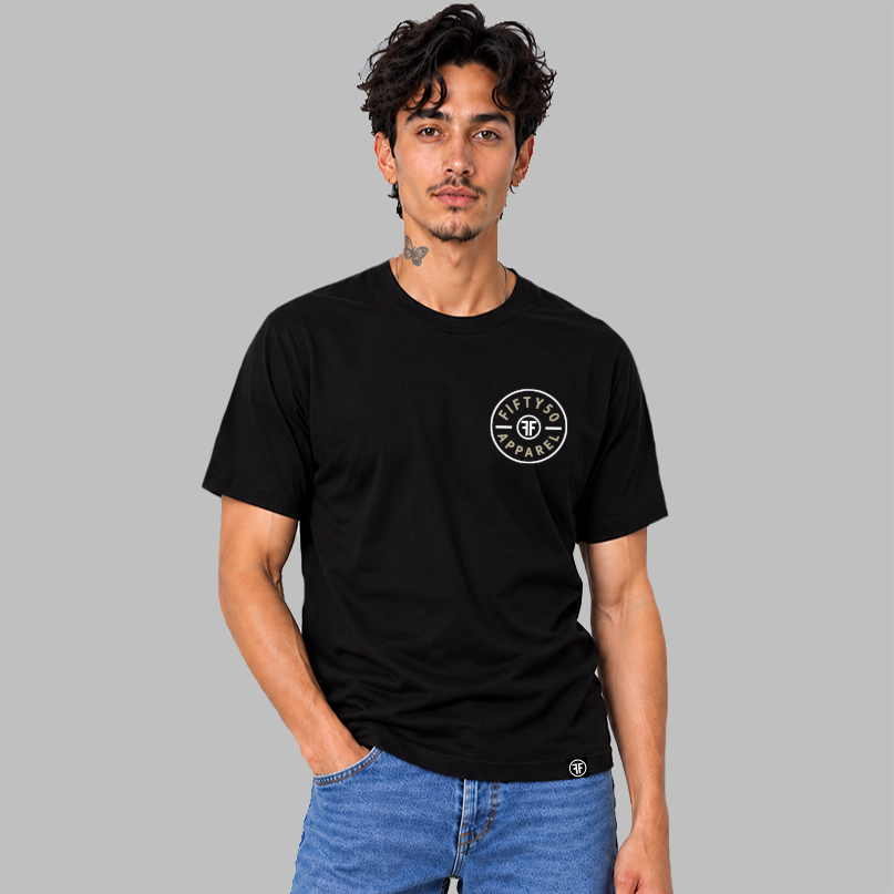 A man wearing a black short-sleeve t-shirt with a circular white chest graphic that reads 'FIFTY50 APPAREL' with two white FFs back to back in the middle, paired with blue jeans, set against a solid gray background.