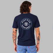 A man viewed from the back wearing  a navy blue crewneck t-shirt with a circular white graphic that says 'FIFTY50 APPAREL' with two Fs back to back on the large center back, paired with light blue jeans, standing against a solid gray studio background.