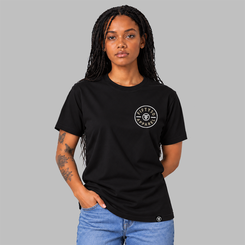 A woman wearing a black short-sleeve t-shirt with a circular white chest graphic that reads 'FIFTY50 APPAREL' with two white FFs back to back in the middle, paired with blue jeans, set against a solid gray background.