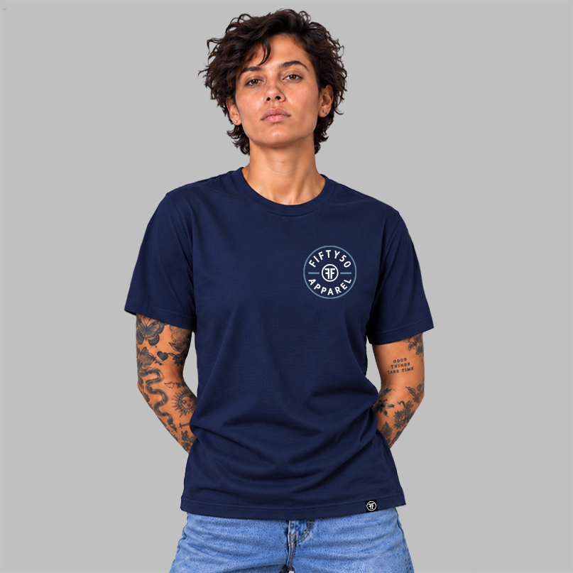 A woman wearing a navy blue crewneck t-shirt with a circular white graphic that says 'FIFTY50 APPAREL' with two Fs back to back on the left chest, paired with light blue jeans, standing against a solid gray studio background.
