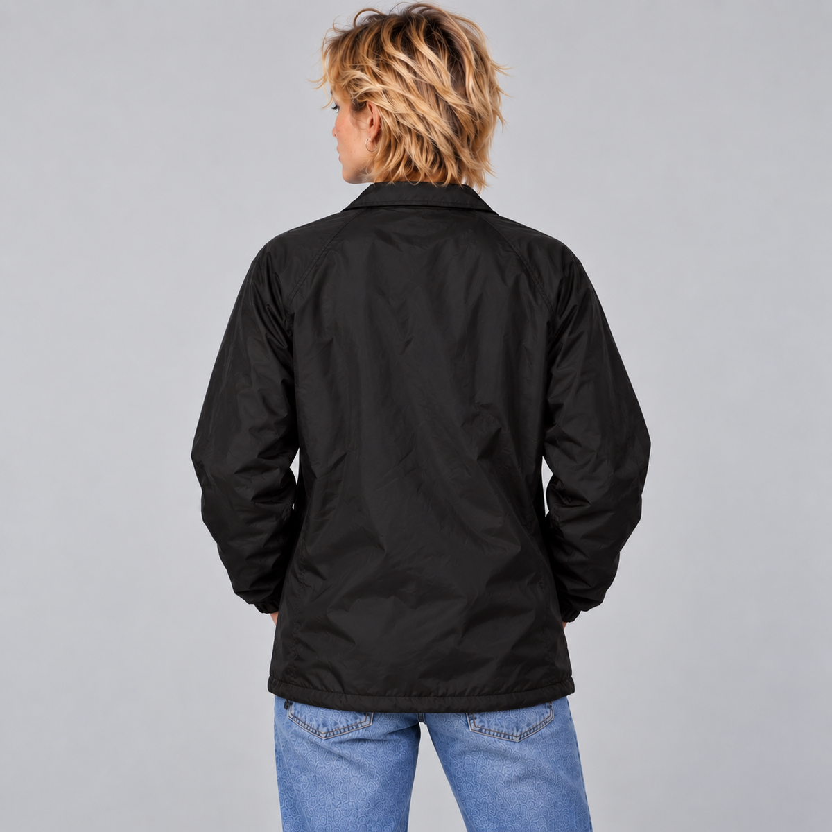 A woman shown from the back wearing a black nylon jacket with a collar and blue jeans against a plain light gray studio background.