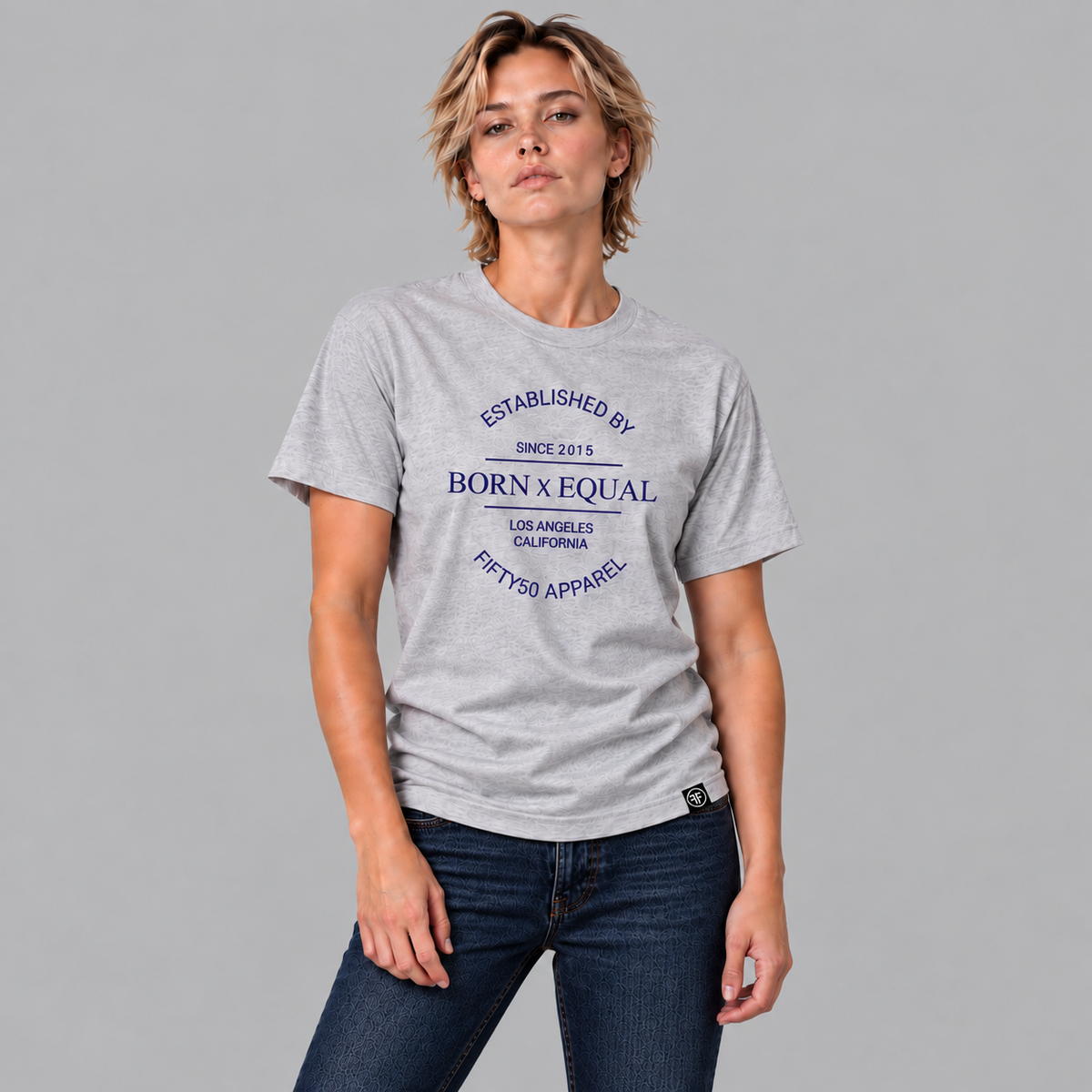 A woman modeling a light heather gray graphic t-shirt with navy blue text that reads 'ESTABLISHED BY SINCE 2015 BORN x EQUAL LOS ANGELES CALIFORNIA FIFTY5O APPAREL', paired with dark blue jeans, against a solid gray background.