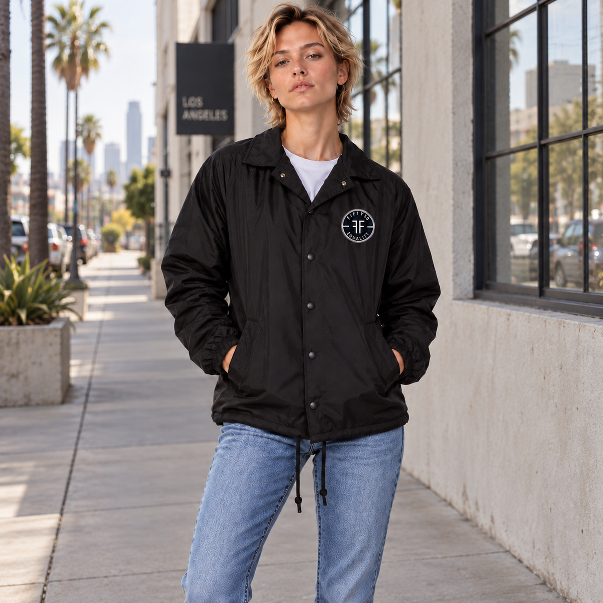 A woman stands on a city sidewalk wearing a black lightweight coach's jacket with a circular black patch left chest that has two FFs in the middle, surrounded by 'Fifty50 Apparel' and 'Equality'. The jacket is paired with a white T-shirt and light blue jeans.
