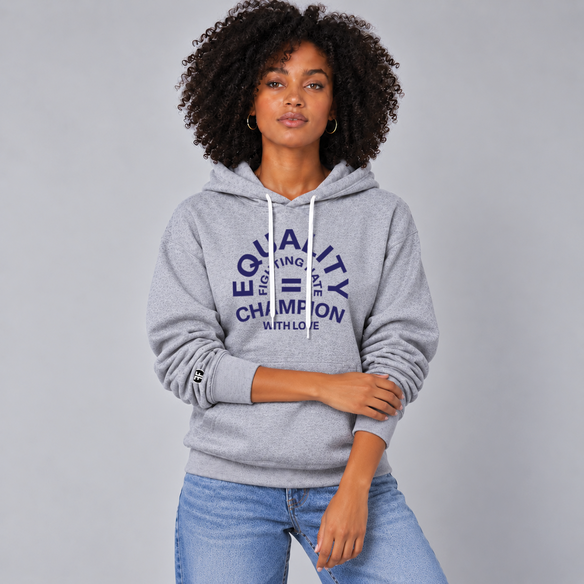 A woman wearing a light gray pullover hoodie with a navy blue graphic on the chest that reads 'EQUALITY CHAMPION FIGHTING HATE WITH LOVE' and a small black circular logo on the left lower sleeve, posed against a solid gray background.