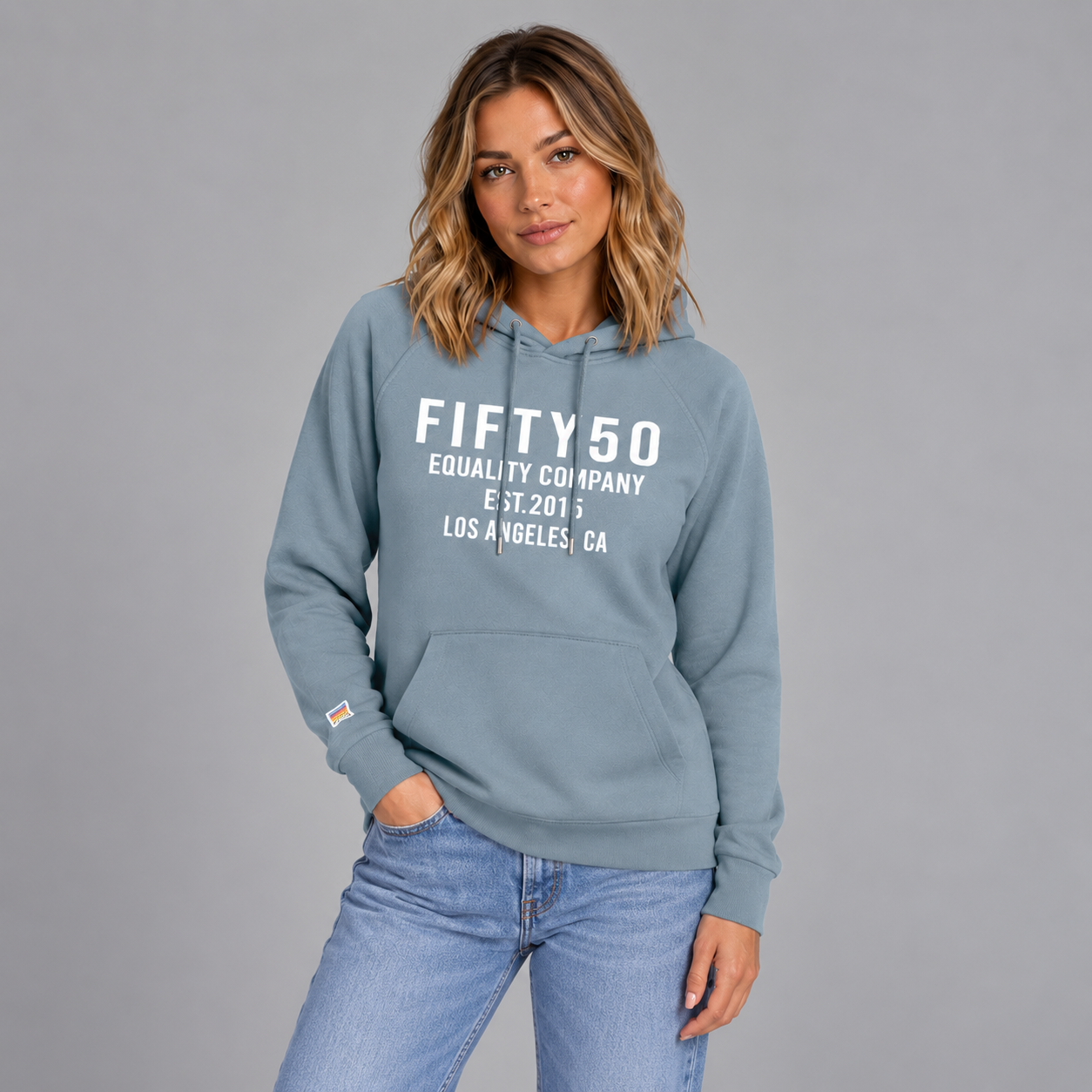 A woman wearing a slate blue pullover hoodie with white text reading 'FIFTY50 EQUALITY COMPANY EST. 2015 LOS ANGELES, CA' on the front, paired with black pants, against a solid gray studio background.