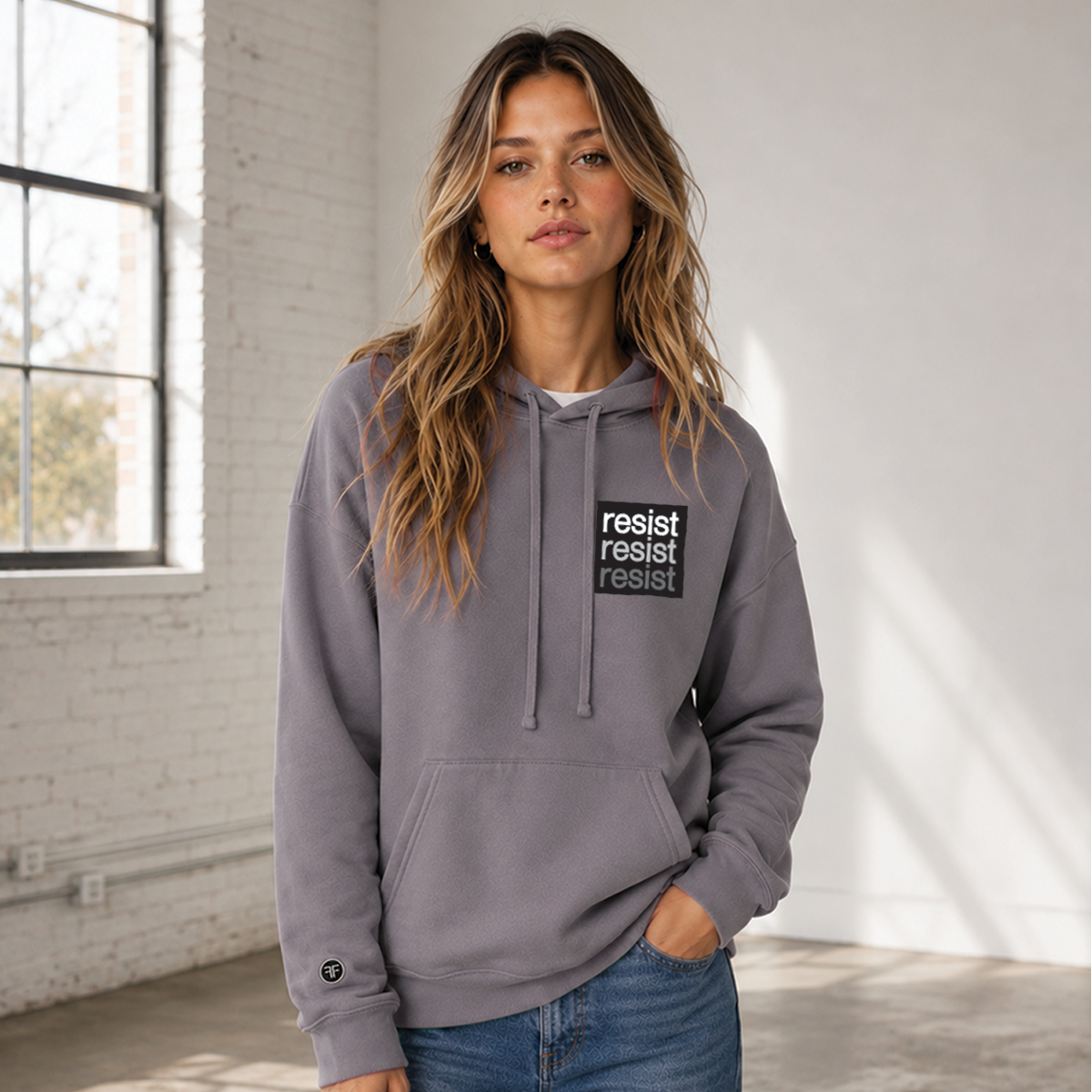 A woman wearing a dusty purple-gray pullover hoodie with a black chest logo that reads 'resist resist resist' in white, light grey and dark grey text, standing in a bright, modern room with white brick walls and a window.