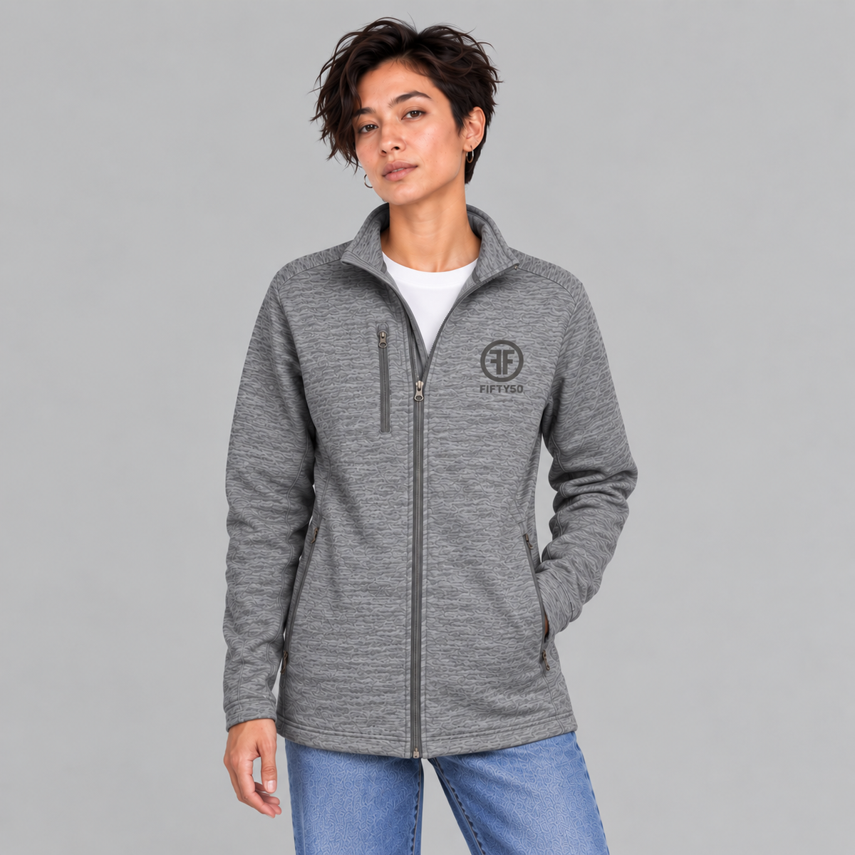 A woman modeling a heather gray full-zip fleece jacket with a stand collar and a circular 'FF' logo on the left chest, paired with a white t-shirt and blue jeans against a solid gray studio background.