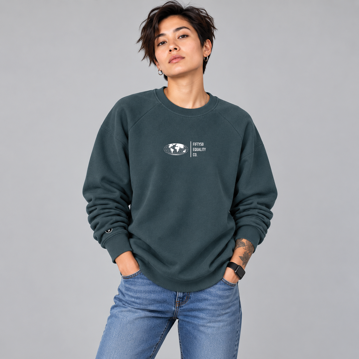 A woman wearing a slate teal crewneck sweatshirt with a small white graphic on the front center chest . The graphic features a globe and the words 'FIFTY50 EQUALITY CO.'
