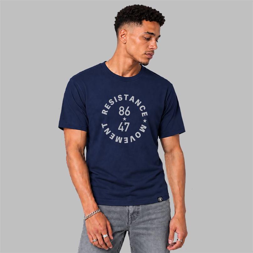 A man wearing a navy blue crewneck t-shirt with a light grey circular graphic that reads 'RESISTANCE MOVEMENT 86 47' on the chest, paired with gray jeans, set against a solid gray background.