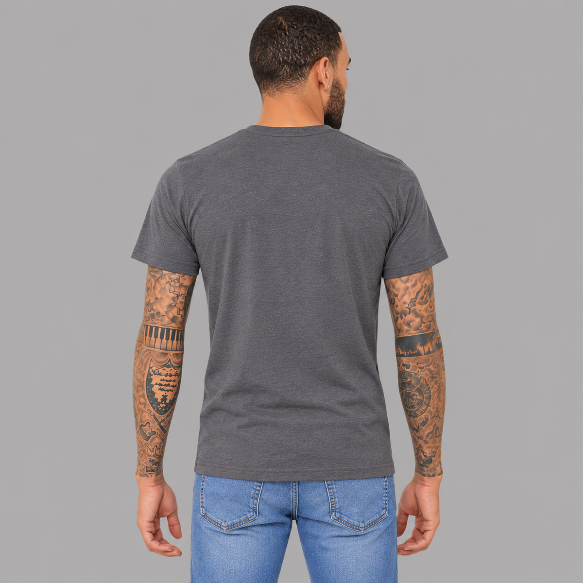 A man viewed from the back wearing a dark heather gray crewneck t-shirt showing a unisex fit and that there are no graphics, and it is paired with blue jeans, standing against a solid gray studio background.