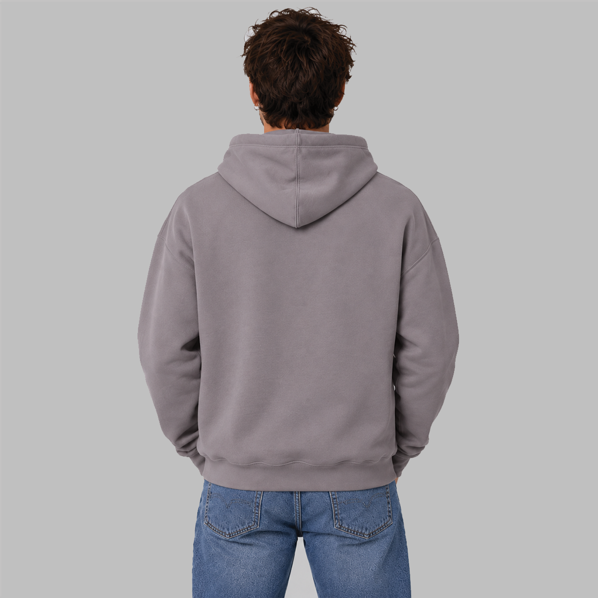 A man viewed from behind wearing a plain slate purple pullover hoodie and blue jeans against a solid gray studio background.