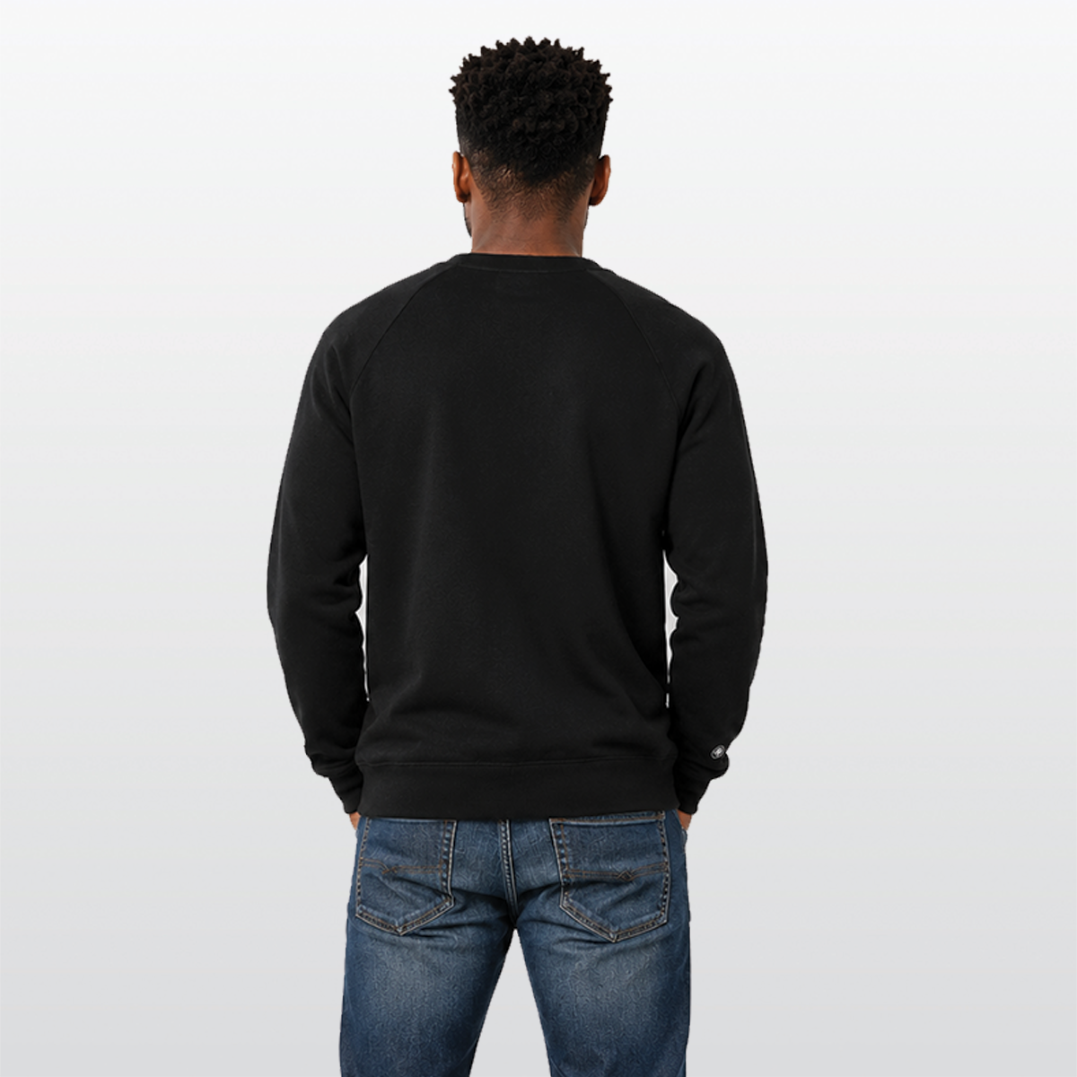 A man viewed from behind wearing a black crewneck sweatshirt and blue jeans, standing against a light gray studio background.