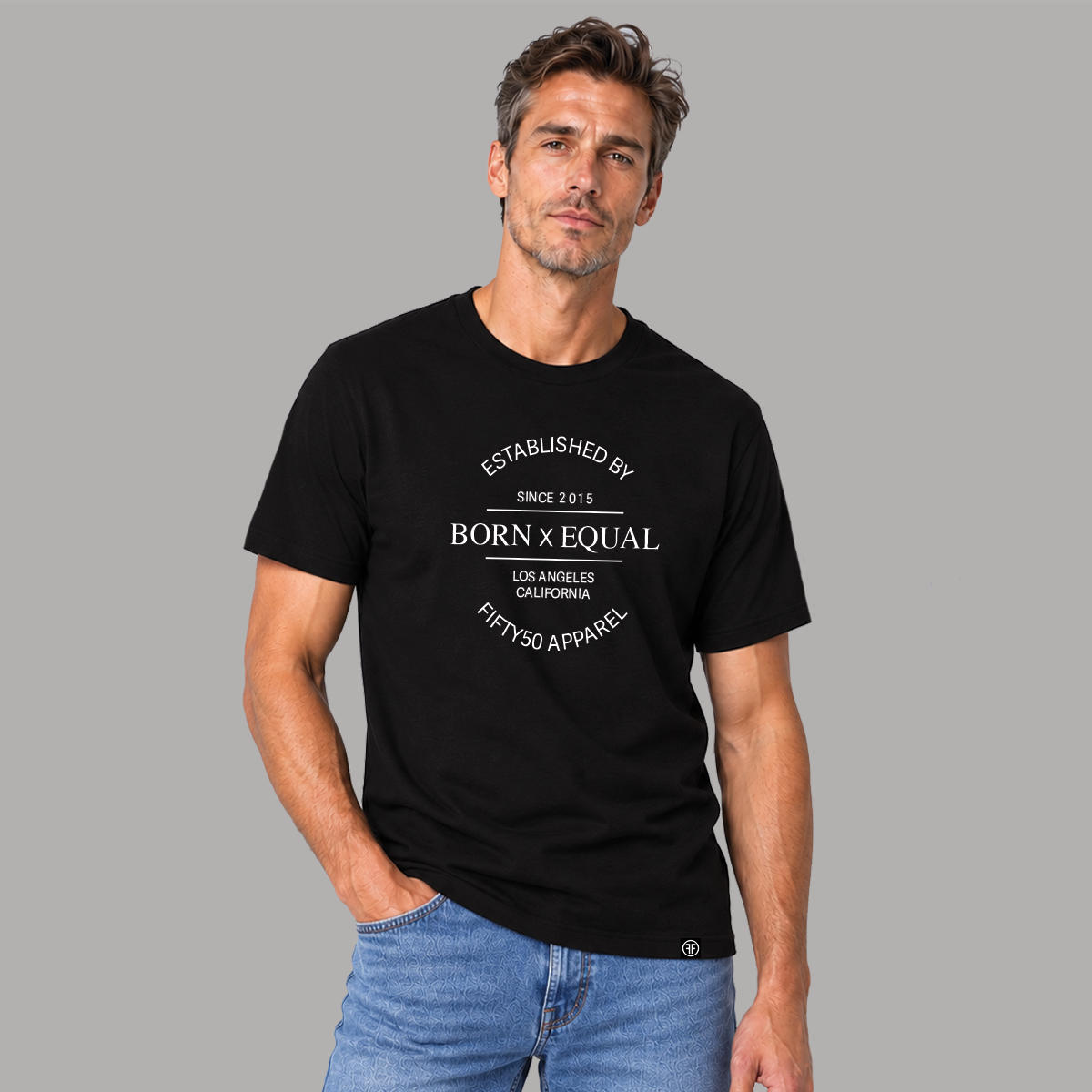 A man with curly black hair models a black short-sleeve graphic t-shirt with white text that reads 'ESTABLISHED BY BORN x EQUAL LOS ANGELES CALIFORNIA FIFTYSO APPAREL'. She is wearing light wash blue jeans against a solid gray studio background.