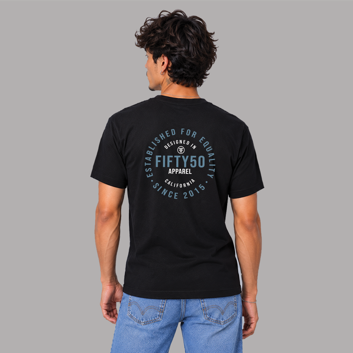 A man viewed from the back wearing a black short-sleeve T-shirt with a circular light blue and white graphic on the back that reads 'ESTABLISHED FOR EQUALITY DESIGNED IN CALIFORNIA,  FIFTY50 APPAREL, SINCE 2015'.