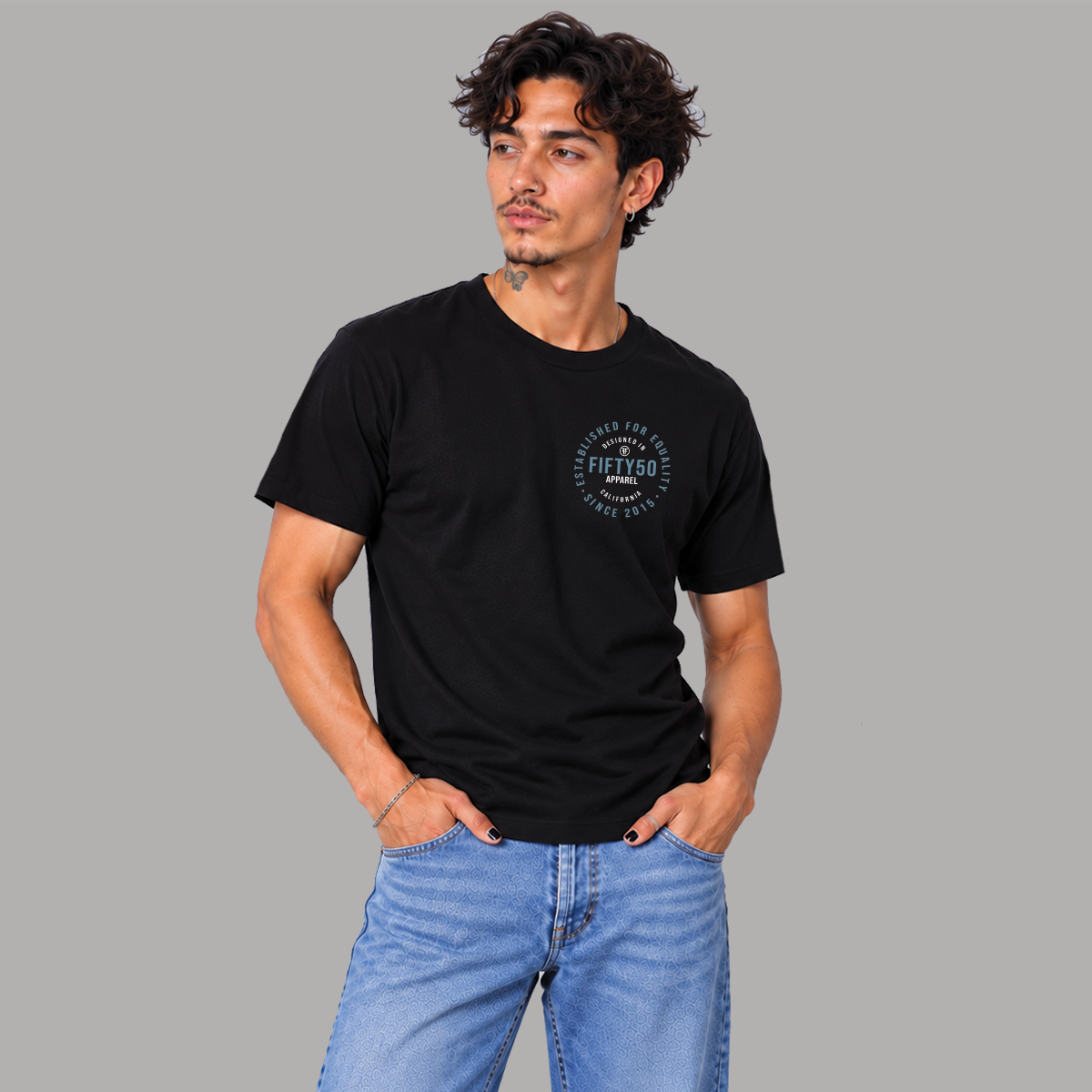 A man wearing a black crew-neck t-shirt with a circular graphic on the left chest, that says 'DESIGNED IN CALIFORNIA, ESTABLISHED FOR EQUALITY, FIFTY50 APPAREL' paired with light blue jeans, standing against a solid gray background.
