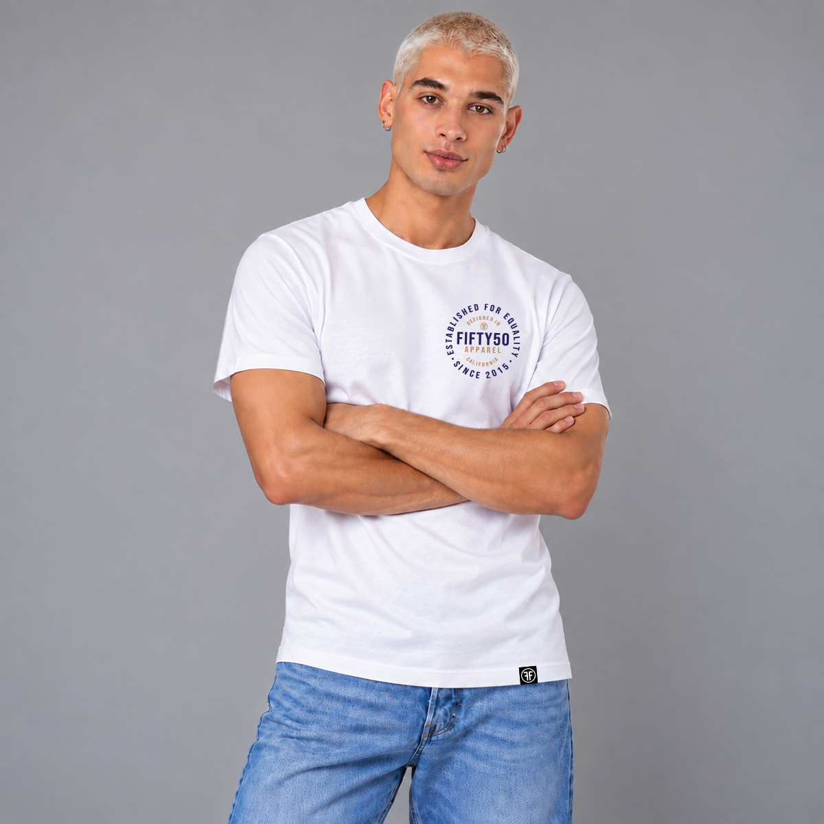 A man wearing a white short-sleeve T-shirt with a circular navy and soft orange graphic print on the left chest reading 'FIFTY50 APPAREL ESTABLISHED FOR EQUITY DESIGNED IN CALIFORNIA SINCE 2015, paired with blue jeans against a solid gray studio background.