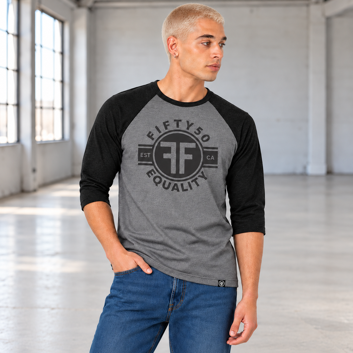 A man wearing a heather grey equality baseball t-shirt with heather black arms and collar. The text of the black graphic says Fifty50, Equality, and has two Fs, back to back in a circle.