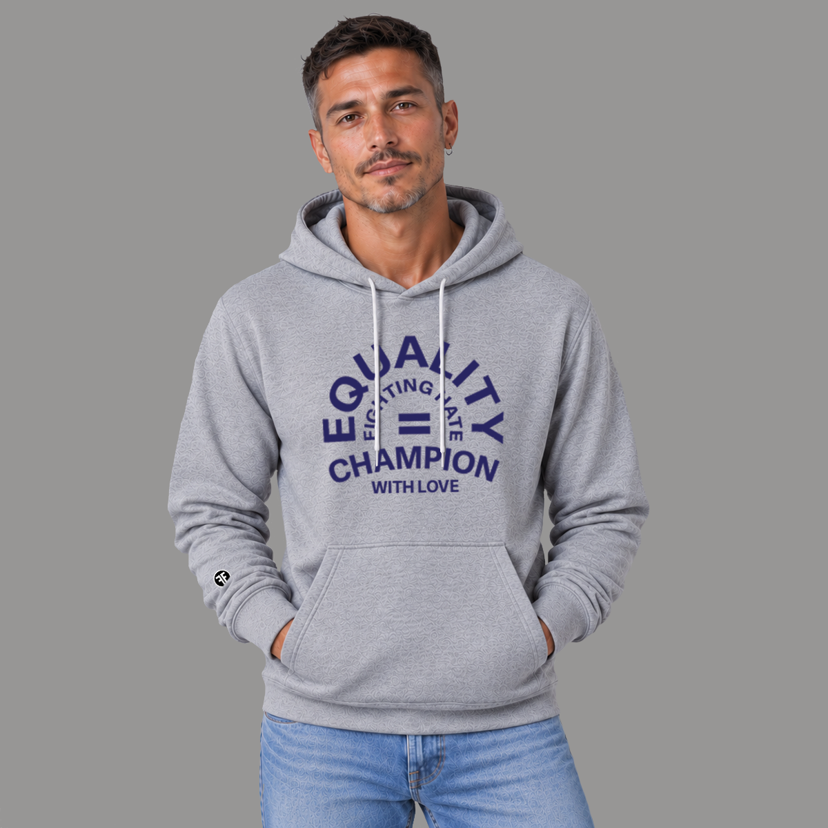 A man wearing a light gray pullover hoodie with a navy blue graphic on the chest that reads 'EQUALITY CHAMPION FIGHTING HATE WITH LOVE' and a small black circular logo on the left sleeve, posed against a solid gray background.