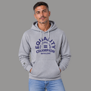 A man wearing a light gray pullover hoodie with a navy blue graphic on the chest that reads 'EQUALITY CHAMPION FIGHTING HATE WITH LOVE' and a small black circular logo on the left sleeve, posed against a solid gray background.