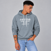 A man wearing a slate blue pullover hoodie with white text reading 'FIFTY50 EQUALITY COMPANY EST 2015 LOS ANGELES, CA' on the chest, paired with light blue jeans, against a solid gray studio background.