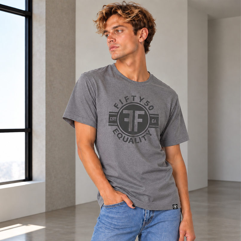 A man wearing a heather gray crewneck t-shirt with a black circular graphic that reads 'FIFTY50 EQUALITY EST. 2025 CA' and light blue jeans.