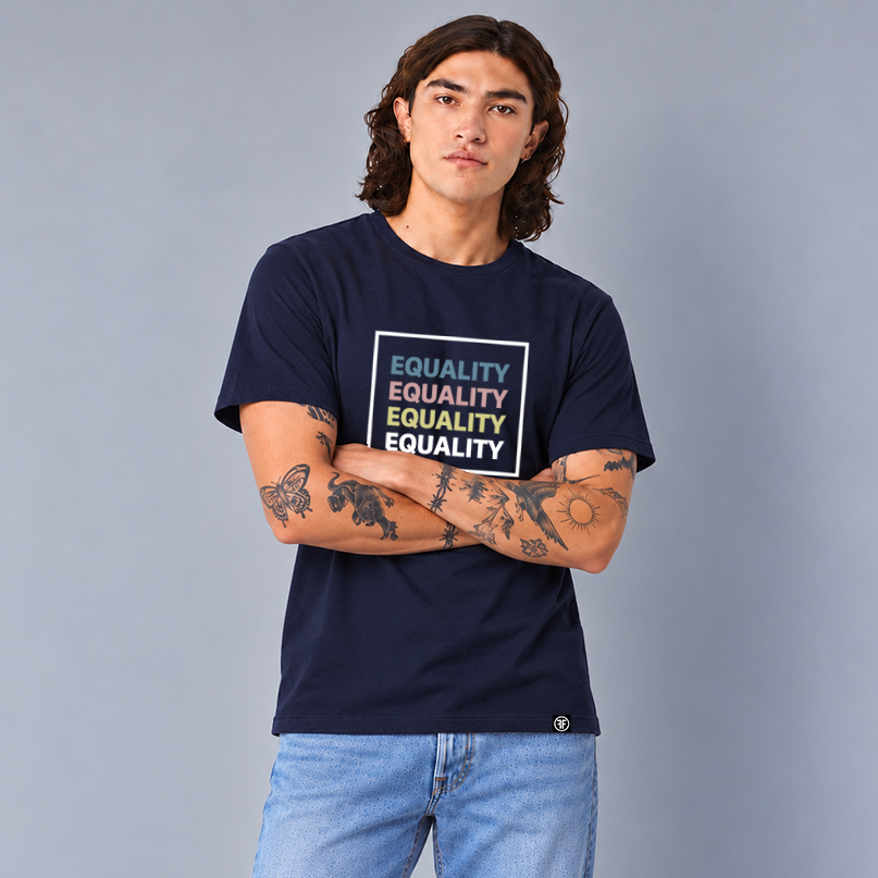 A man with long dark hair and tattoos stands with arms crossed, wearing a navy blue crewneck t-shirt with a colorful graphic on the chest that reads 'EQUALITY' repeated in slate blue, pink, yellow, and white text, paired with light blue jeans, against a solid gray studio background.