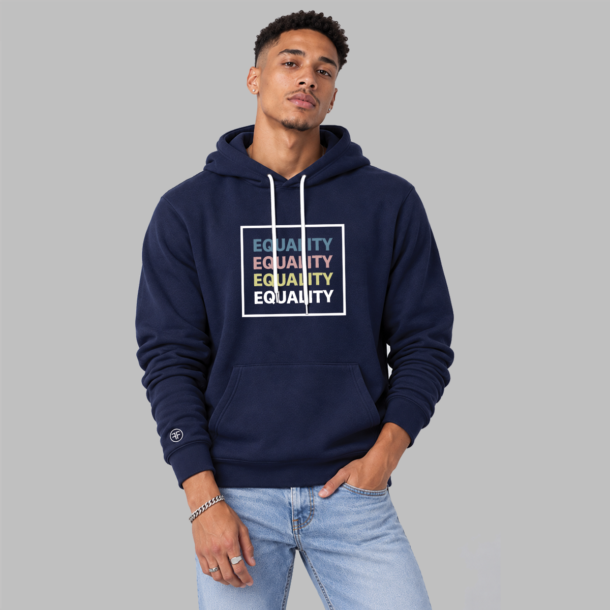A man wearing a navy blue hoodie with a white drawstring and a graphic on the chest that reads 'EQUALITY' repeated in blue, pink, yellow and white text inside a white square. She is also wearing light blue jeans, standing in a minimalist studio with a white wall and gray floor.