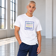 A man wearing a white crewneck t-shirt with a colorful graphic print that reads 'EQUALITY' repeated in stone blue, pink, yellow, and navy blue, paired with navy blue pants sweatpants, standing  a bright, modern studio with large windows.