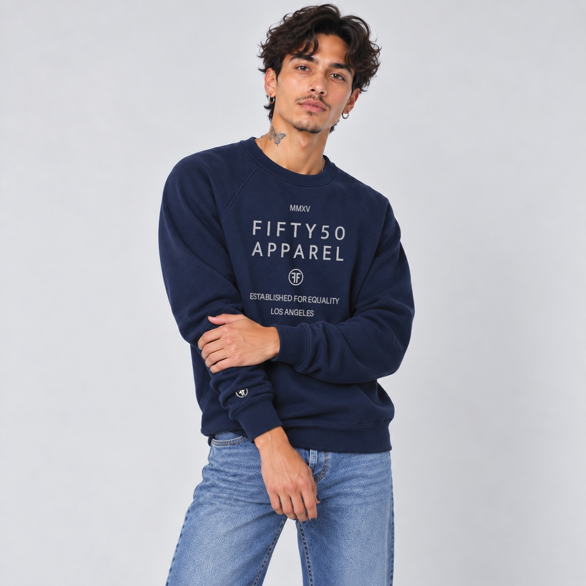 A man wearing a navy blue crewneck sweatshirt with 'FIFTY50 APPAREL ESTABLISHED FOR EQUALITY LOS ANGELES' printed in light grey on the front, paired with light-wash blue jeans, against a light gray studio background.