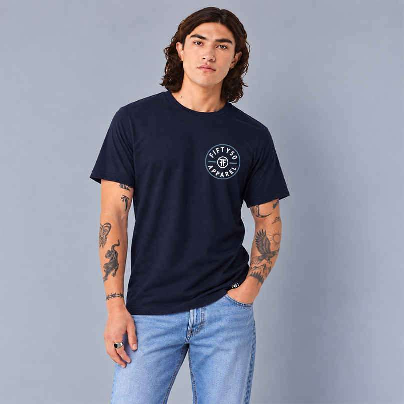 A man wearing a navy blue crewneck t-shirt with a circular white graphic that says 'FIFTY50 APPAREL' with two Fs back to back on the left chest, paired with light blue jeans, standing against a solid gray studio background.