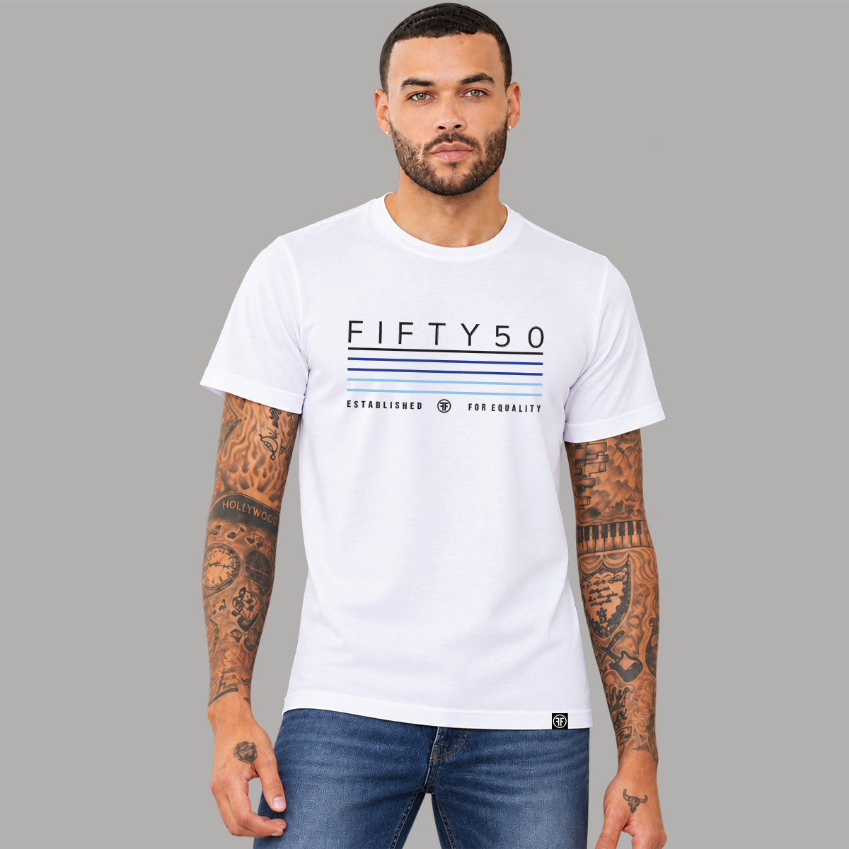 A man wearing a white short-sleeve graphic T-shirt with 'FIFTY50' and thin black, navy, royal, and light blue stripes printed on the front, paired with light blue jeans, against a plain gray studio background.